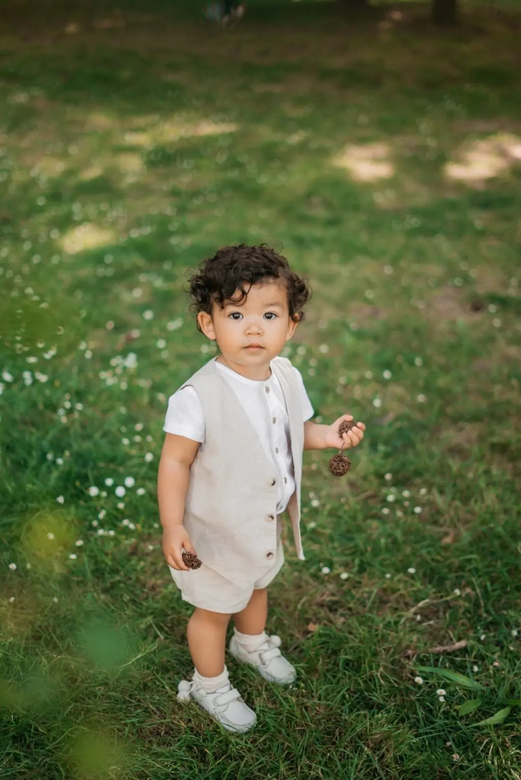 A toddler with dark curls in a linen vest standing on the grass