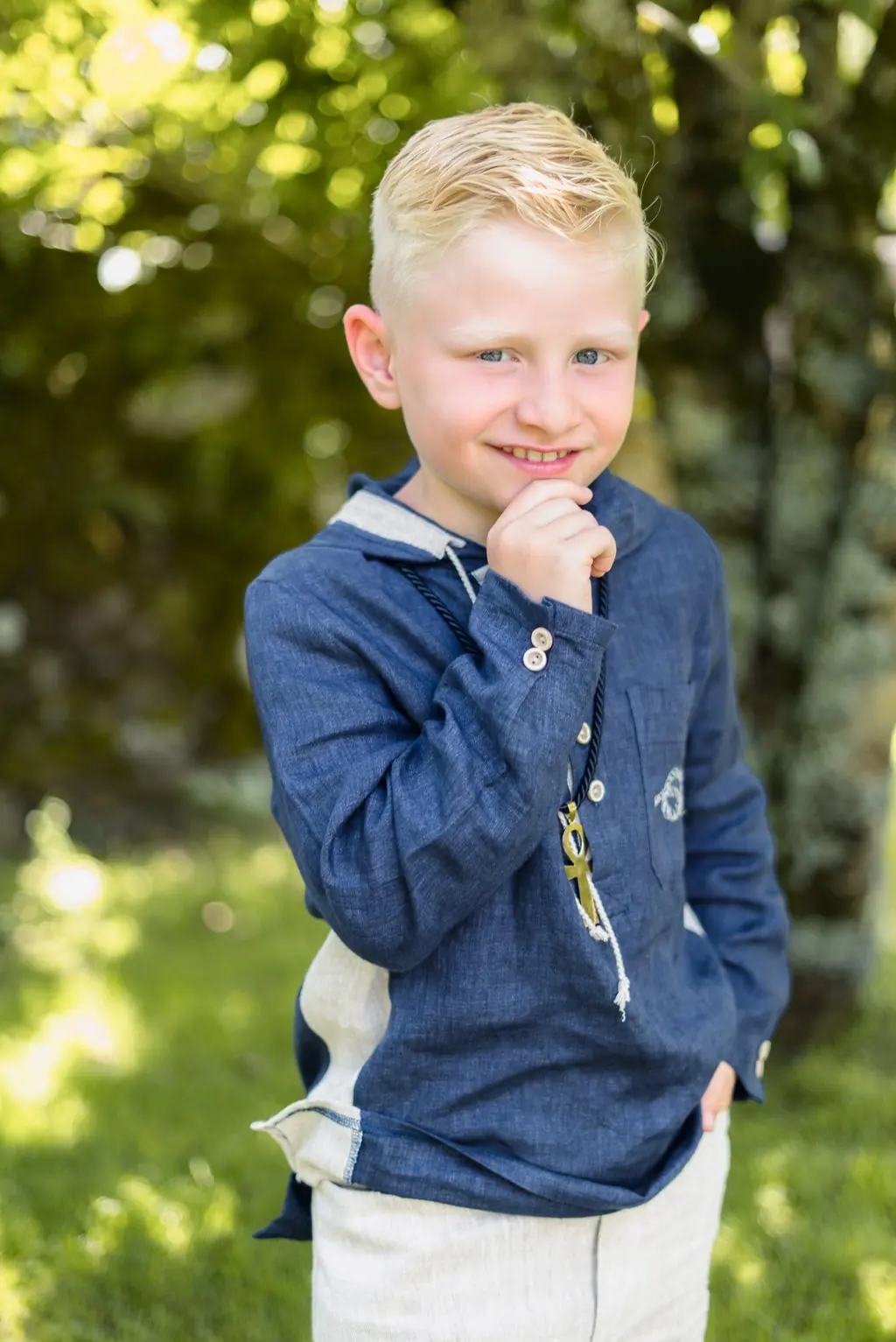 A blond boy in a blue shirt smiles with a hand at his chin