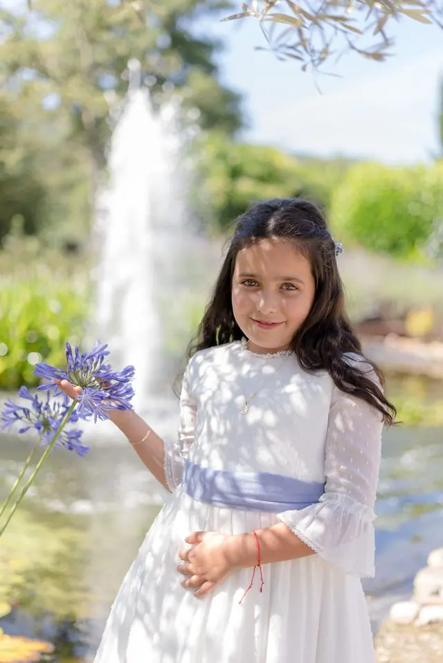 Una niña con vestido blanco y faja azul sostiene una flor morada junto a una fuente