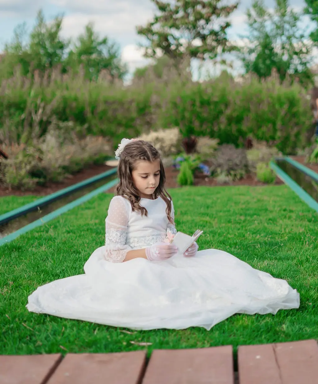A girl in a communion dress seated on the grass holding a small prayer book
