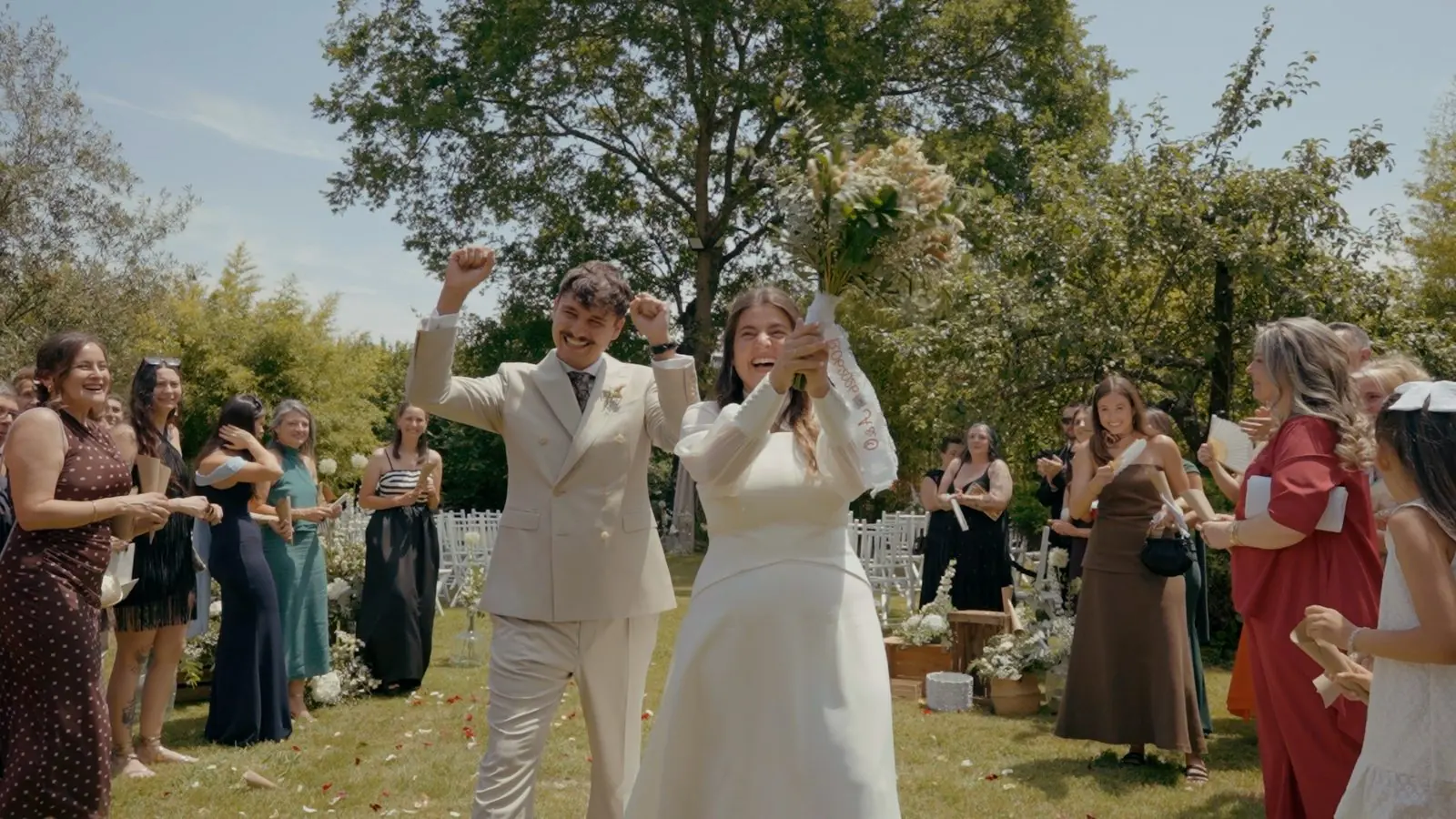 Los novios levantan los brazos, celebrando entre sus invitados en un jardín