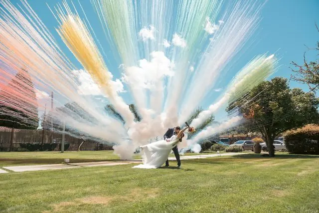Una pareja de novios en un jardín mientras estalla polvo de colores en el aire a su espalda