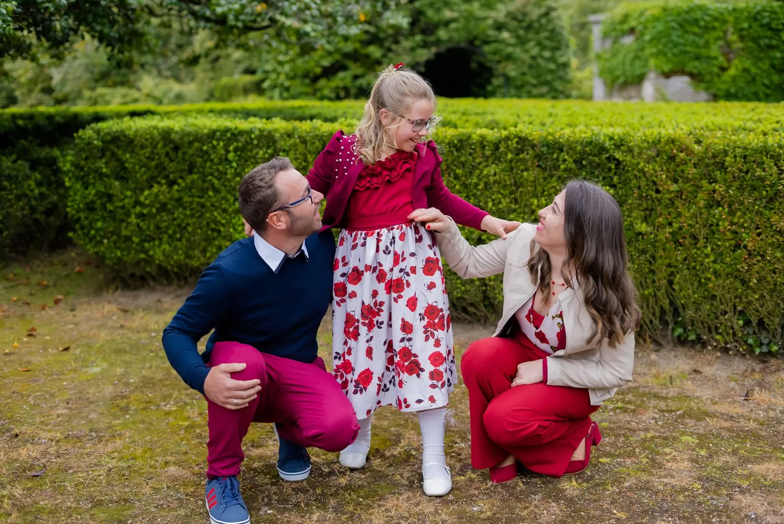 Parents crouch to embrace their daughter in a hedge garden