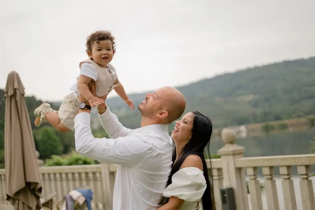 A father lifts his toddler while the mother smiles beside a lake