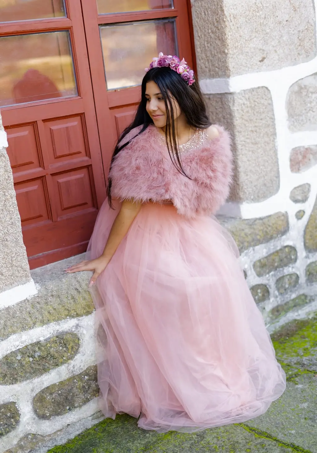A young woman in a pink tulle dress seated by a red door in a stone wall