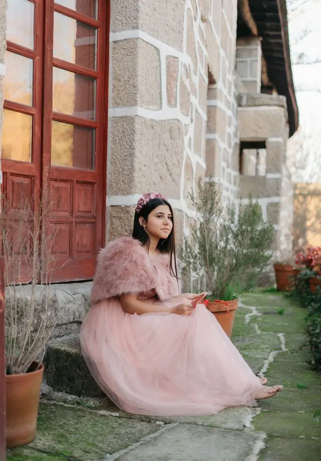 Una joven con vestido rosa sentada en un escalón de piedra, con una puerta roja detrás