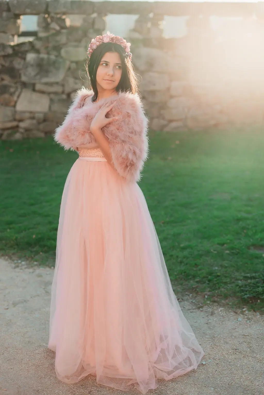 A young woman in a pink tulle skirt and feather bolero, backlit beside a stone wall